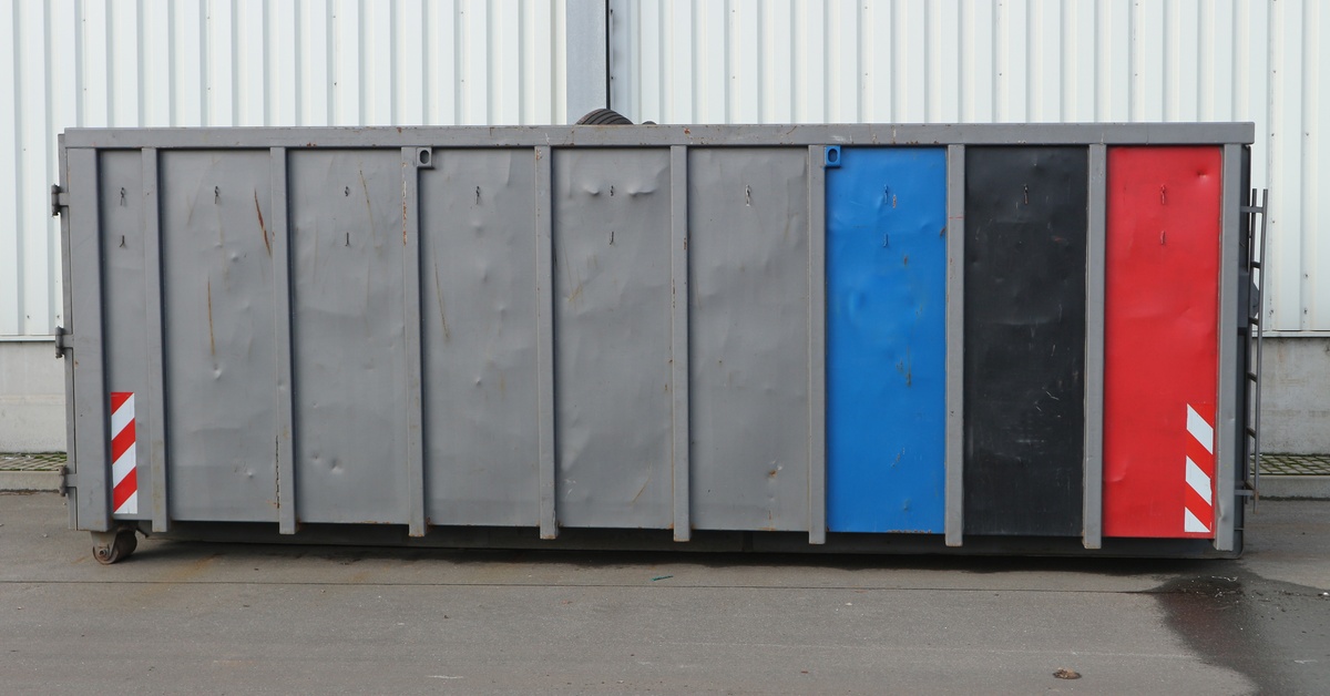 A large grey dumpster with a blue, a black, and a red stripe on the right side, placed on concrete next to a white wall.