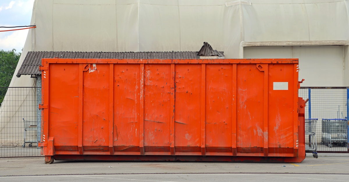 A large orange dumpster placed at the back of a large building with grey walls next to multiple shopping carts.
